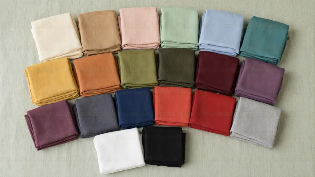 Twenty folded plain hijabs on a linen cloth, from light shades such as cream, blush, mint and light blue to deep ones such as mustard, terracotta, burgundy, plum, navy and black.