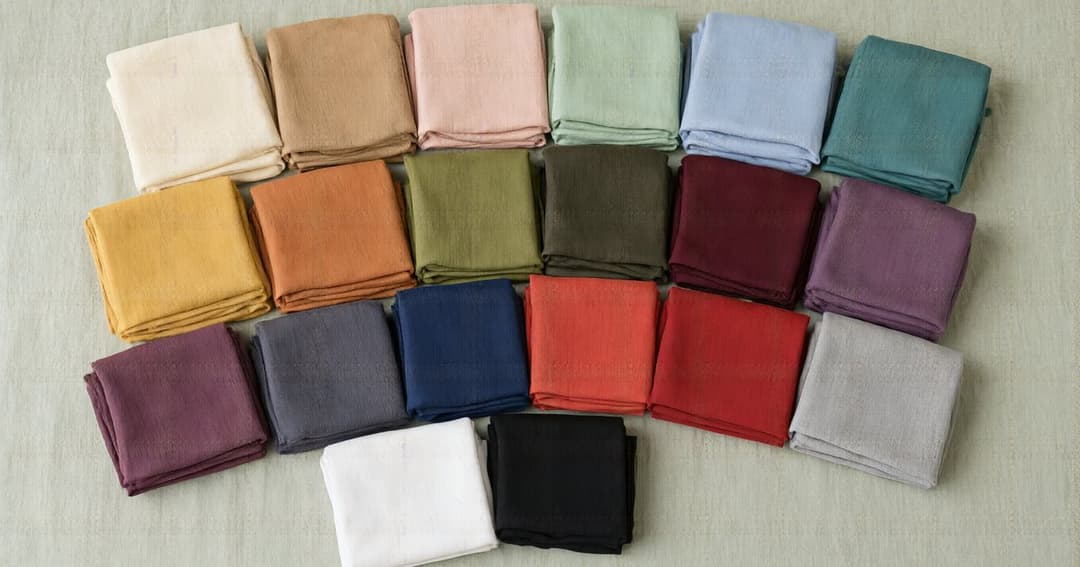 Twenty folded plain hijabs on a linen cloth, from light shades such as cream, blush, mint and light blue to deep ones such as mustard, terracotta, burgundy, plum, navy and black.